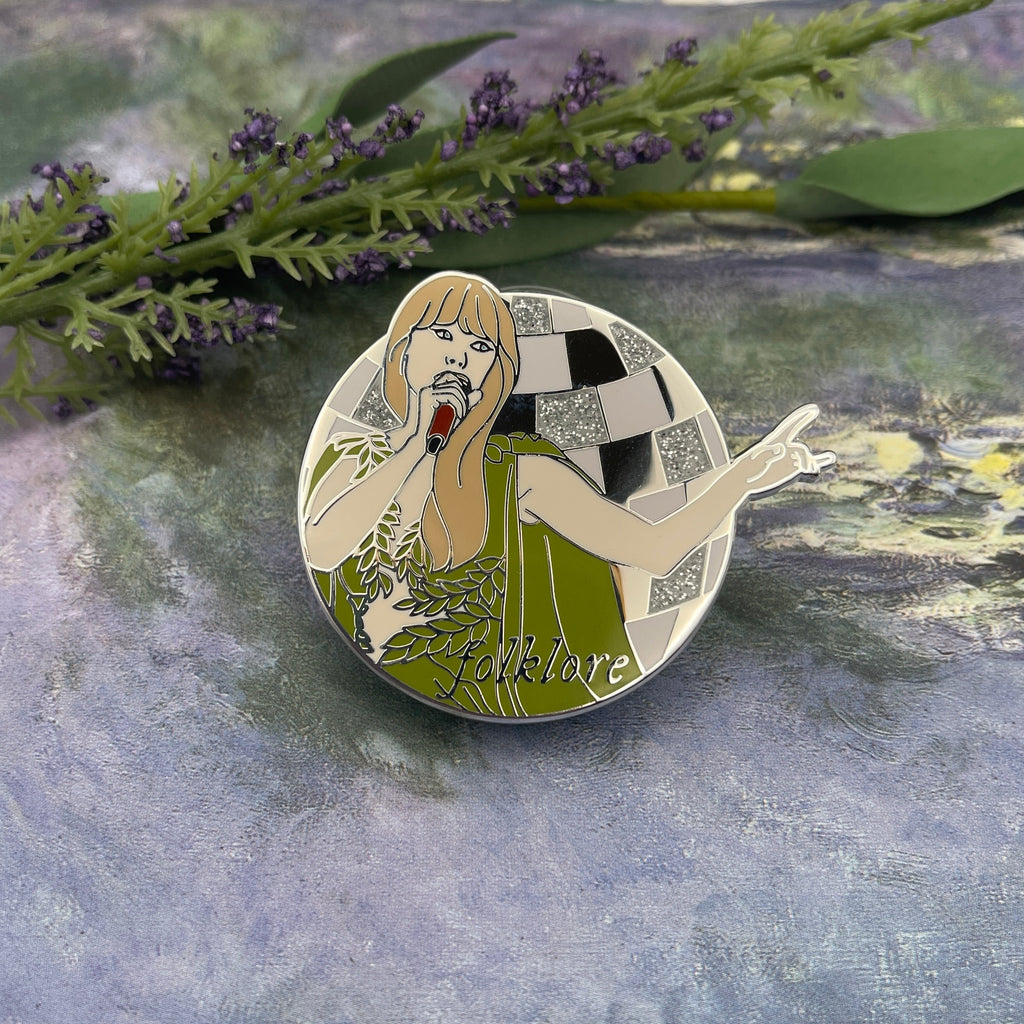 A circular enamel pin featuring an Art Deco style illustration of a woman in a green dress with a checkered background, placed on a textured gray surface with green foliage in the background.
