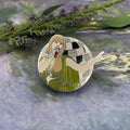 A circular enamel pin featuring an Art Deco style illustration of a woman in a green dress with a checkered background, placed on a textured gray surface with green foliage in the background.