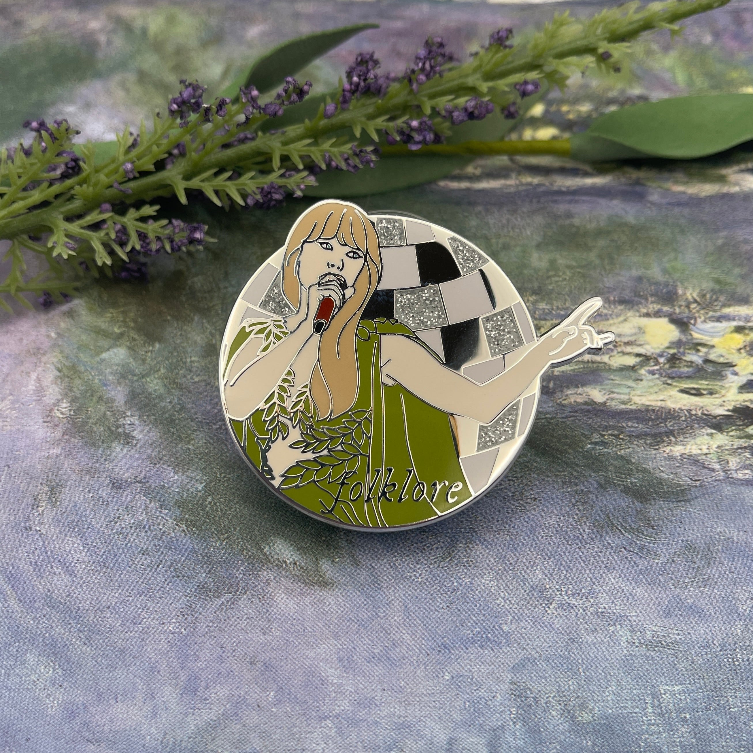 A circular enamel pin featuring an Art Deco style illustration of a woman in a green dress with a checkered background, placed on a textured gray surface with green foliage in the background.